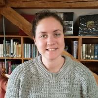 A smiling woman wearing a light sweater stands in front of a book shelf.
