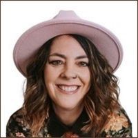 A smiling woman with shoulder length hair and a nose ring wearing a pink hat.