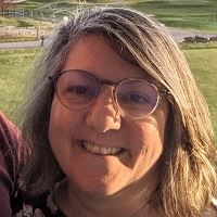 A smiling woman wearing glasses, standing outside in a grassy area.