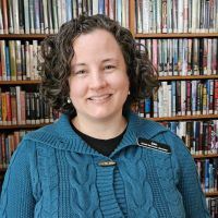 A smiling woman with short dark curly hair, wearing a green cardigan stands in front of bookshelves.