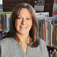 A smiling woman with shoulder length brown hair stands in front of a bookshelf.