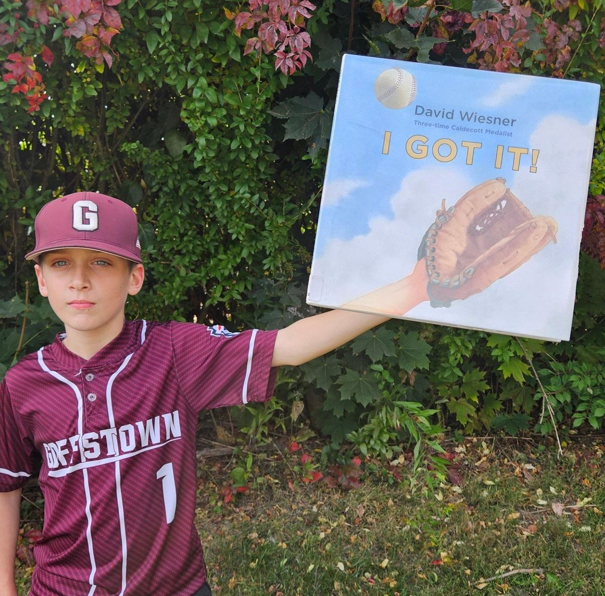 A young man in a Goffstown ball uniform reaches out behind a book with a baseball glove on the cover