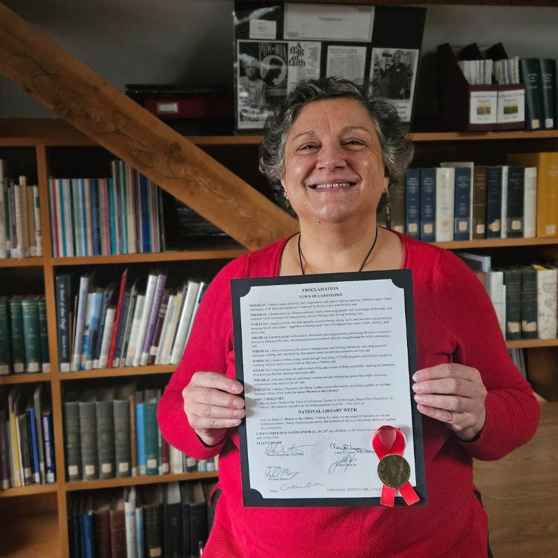 Dianne Hathaway smiles in front of bookshelves, holding a Proclamation from Selectboard.